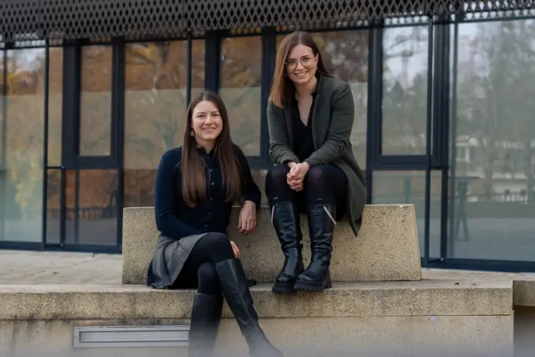 Zwei Frauen sitzen auf einer Betontreppe und lächeln in die Kamera