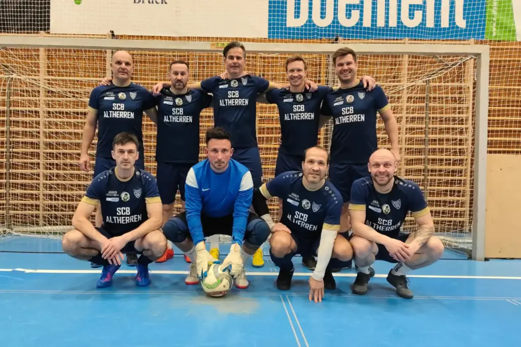 Neun Männer in Fußballdressen vor einem Fußballtor in einer Sporthalle.