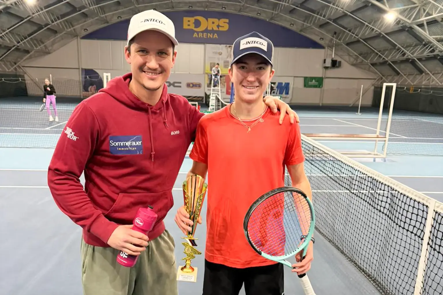 Ein junger Mann mit Trinkflasche in der Hand und ein Jugendlicher mit Tennisschläger und Pokal in der Hand stehen auf einem Tennisplatz.