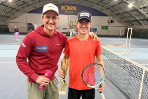Ein junger Mann mit Trinkflasche in der Hand und ein Jugendlicher mit Tennisschläger und Pokal in der Hand stehen auf einem Tennisplatz.