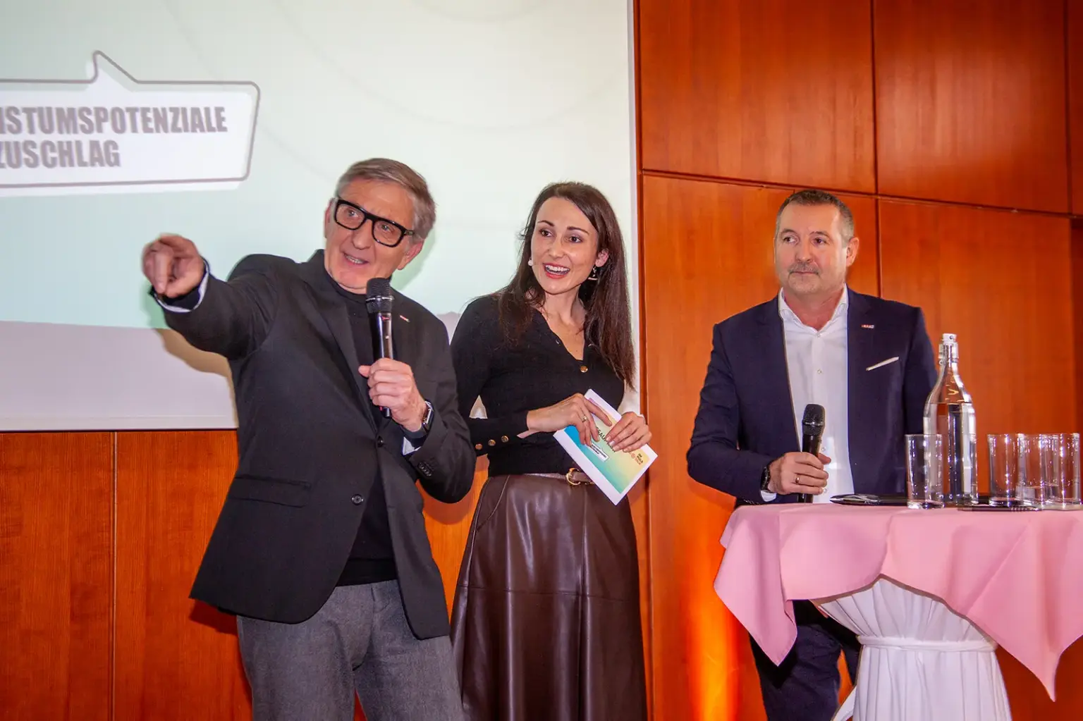 Eine Frau und zwei Männer mit Mikrofonen auf einer Bühne.