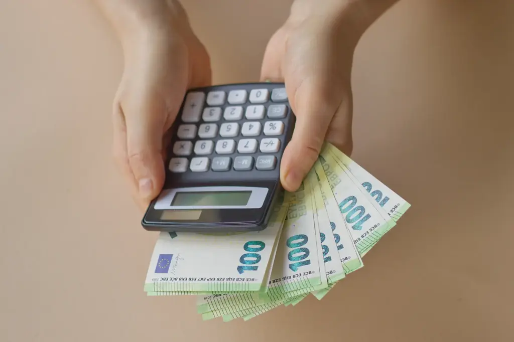 Zwei Hände mit einem Taschenrechner sowie 100-Euro-Scheine dahinter.