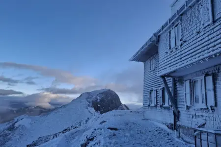 Ein stark mit Raureif bedecktes Haus, dahinter ein Berggipfel mit Kreuz