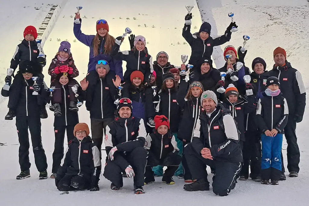 Eine Gruppe von Menschen mit Pokalen und Medaillen vor einer Schisprungschanze.