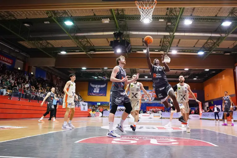 Eine große Sporthalle mit einer Tribüne links, im Mittelpunkt sind Basketballer. Einer davon versucht gerade den Ball in den Korb zu schießen.