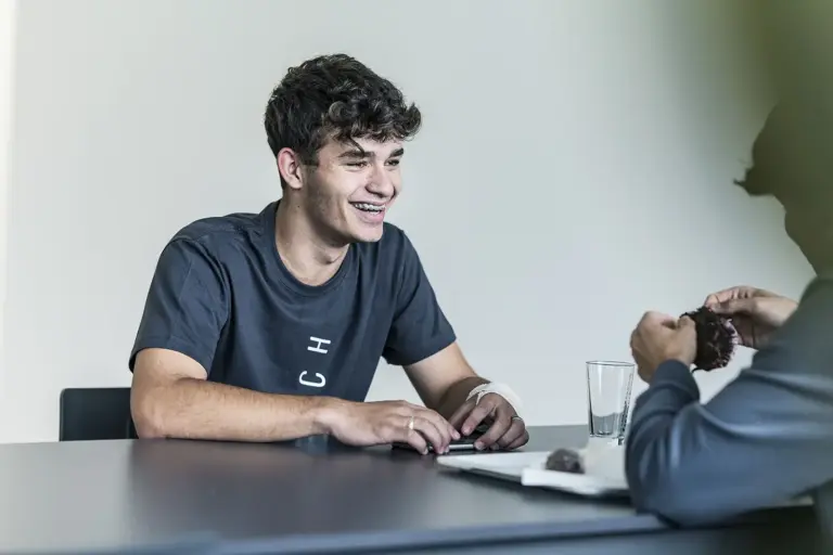 Ein junger Mann sitzt bei einem Tisch und führt ein Gespräch mit einer anderen Person, die nur unscharf am Rand erkennbar ist.
