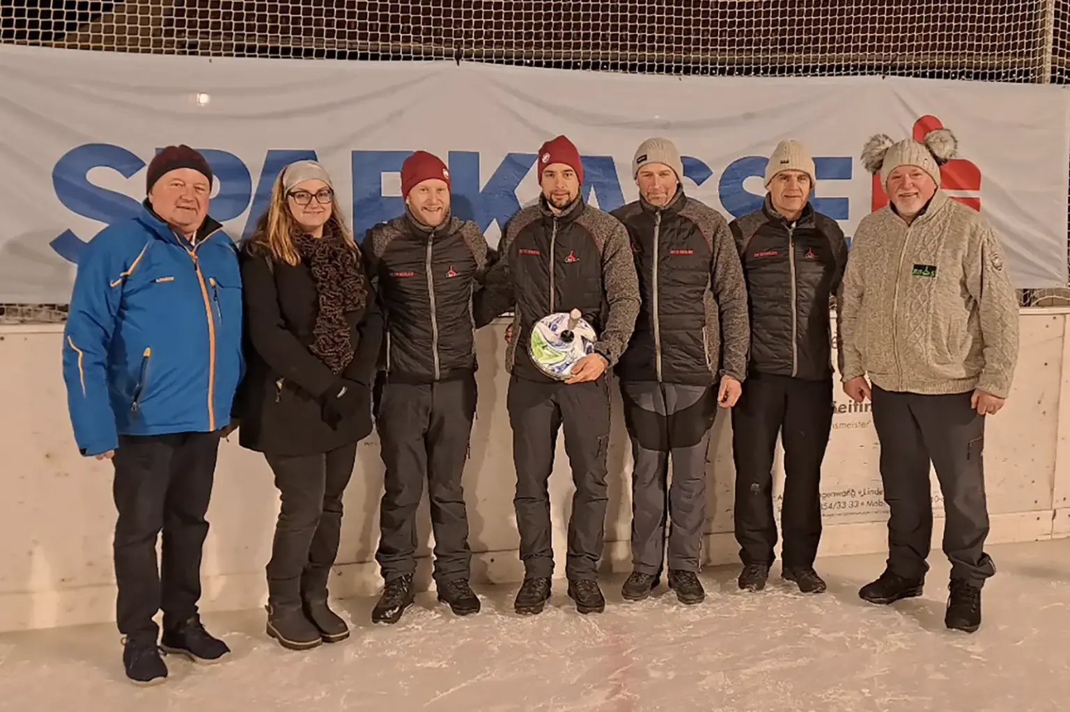Sechs Herren und eine Dame auf der Eisbahn, ein Mann in der Mitte hält einen Eisstock in der Hand.