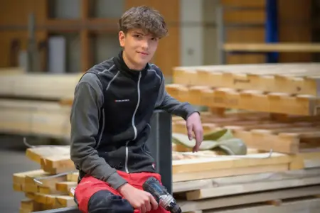 Ein junger Mann mit einem Akkuschrauber in der Hand, er sitzt auf Holzblöcken.