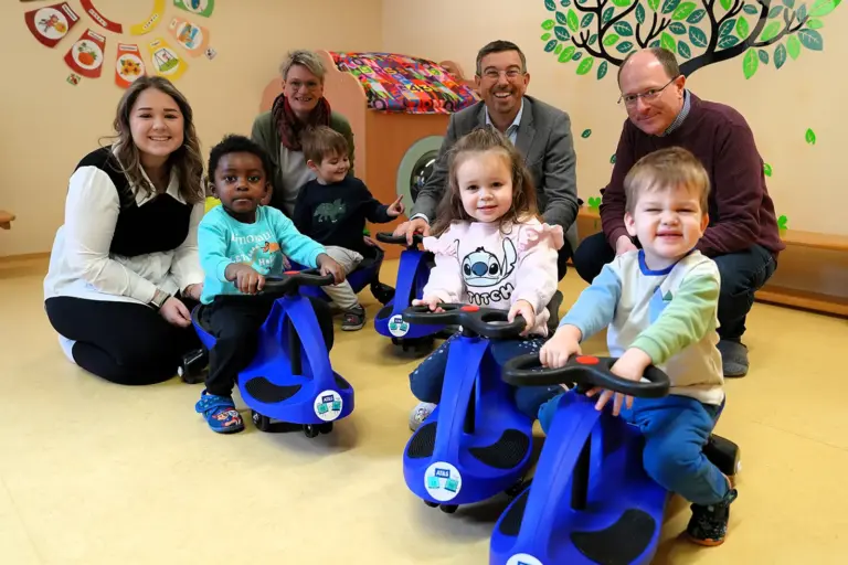 Vier Erwachsene hocken im Kindergarten am Boden. Davor vier Kinder auf blauen Bobbycars.