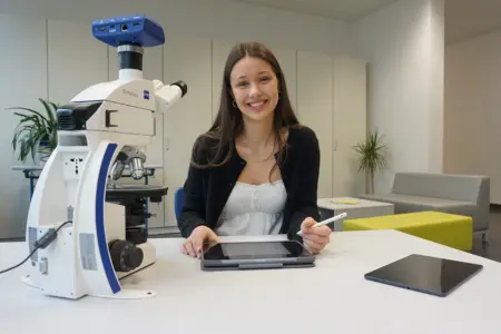 Eine junge Dame hat ein Tablet in der Hand, auf dem Tisch vor ihr steht ein Mikroskop.