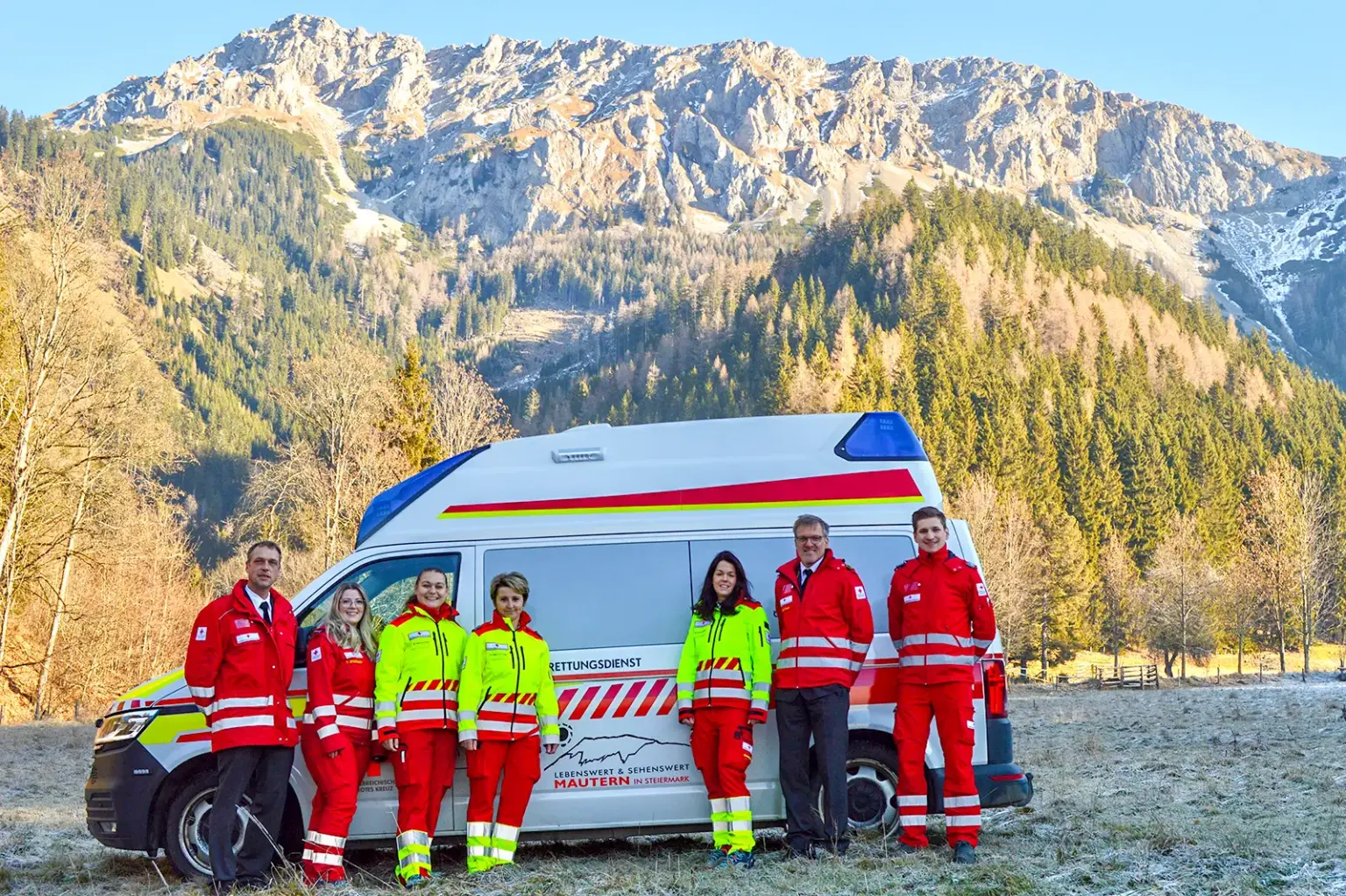 Vier Frauen und drei Männer in Rotkreuz-Dienstkleidung mit Rettungsauto vor Bergkulisse