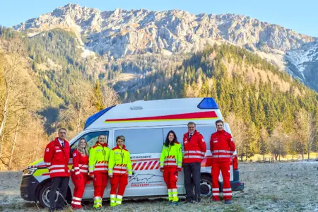 Vier Frauen und drei Männer in Rotkreuz-Dienstkleidung mit Rettungsauto vor Bergkulisse