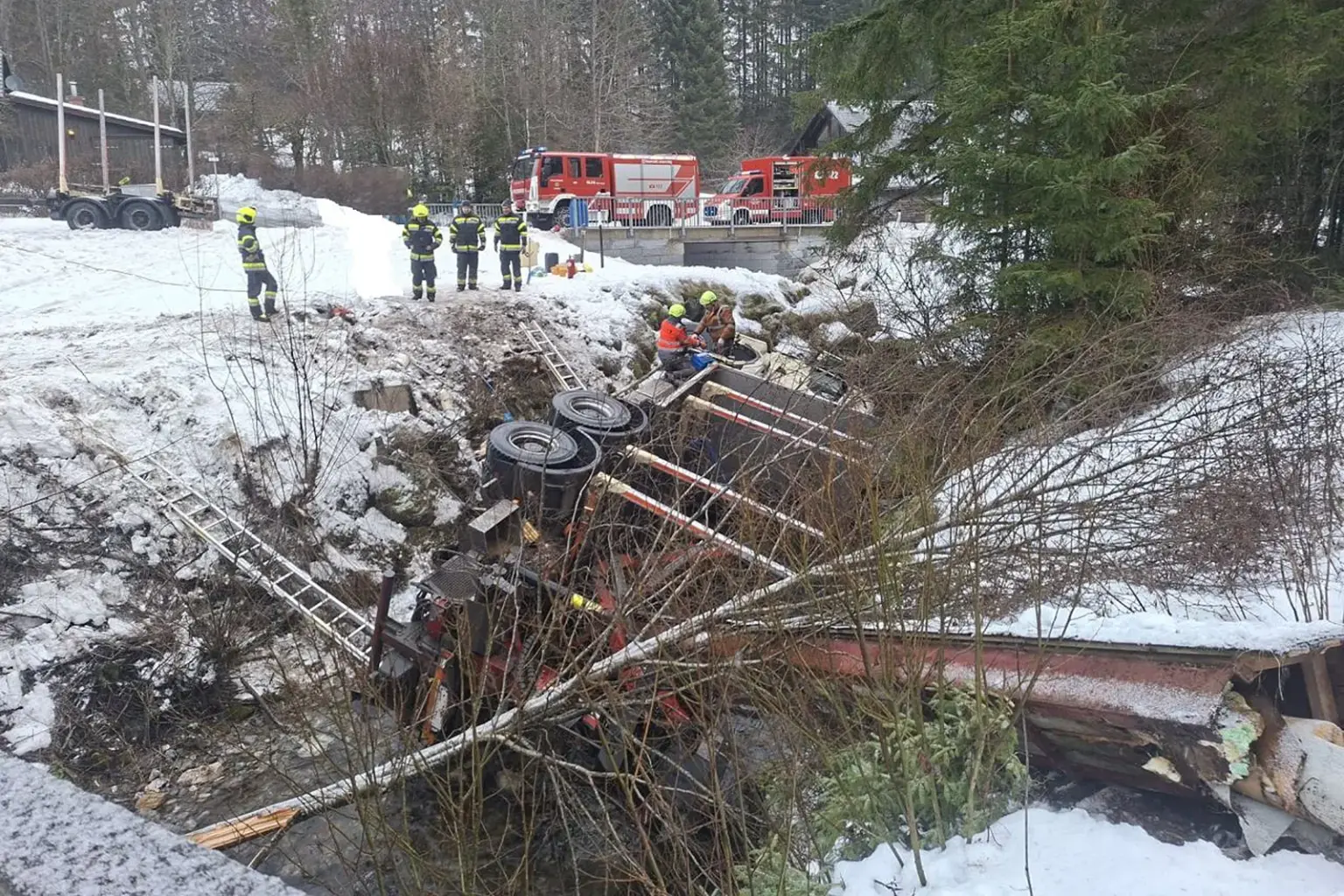 Ein LKW liegt in einem Bach, daneben sind Feuerwehrmänner zu erkennen, die helfen.