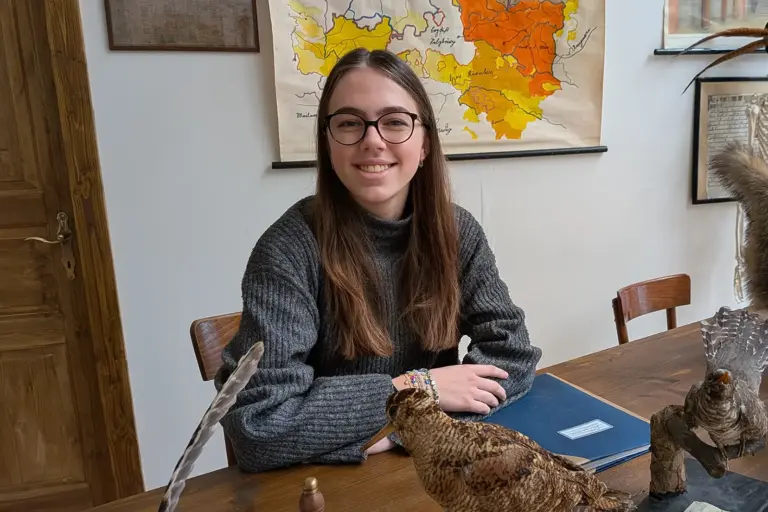 Eine junge Damen sitzt bei einem Tisch und lächelt in die Kamera.