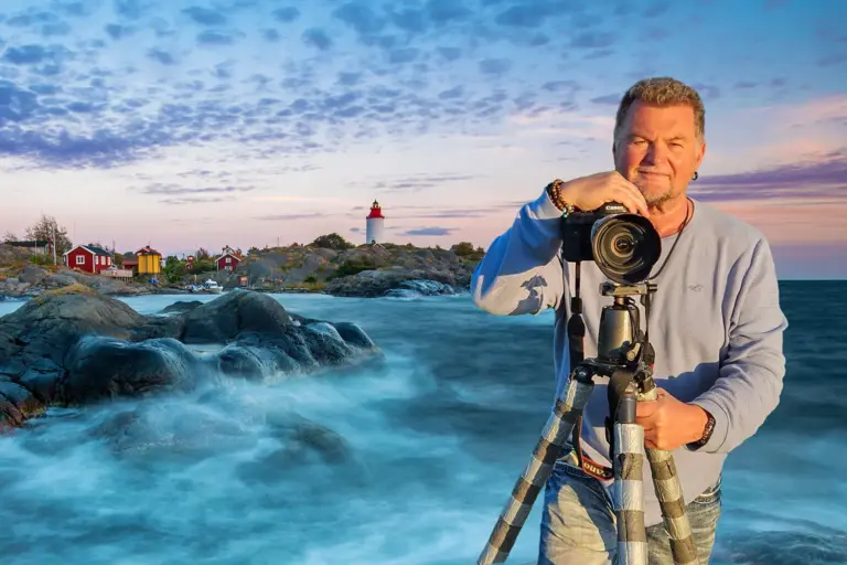 Ein Mann mit einer Kamera auf einem Stativ steht vor einer mystischen Landschaft mit Meer, Leuchtturm und kleinen Häusern