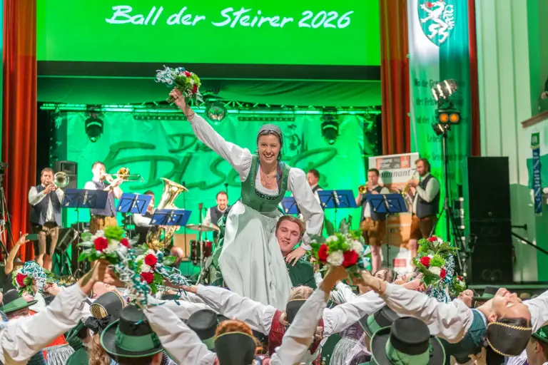 Eine Frau in Tracht sitzt auf der Schulter eines Mannes und winkt mit einem Blumenstrauß, ringsum eine Tanzgruppe
