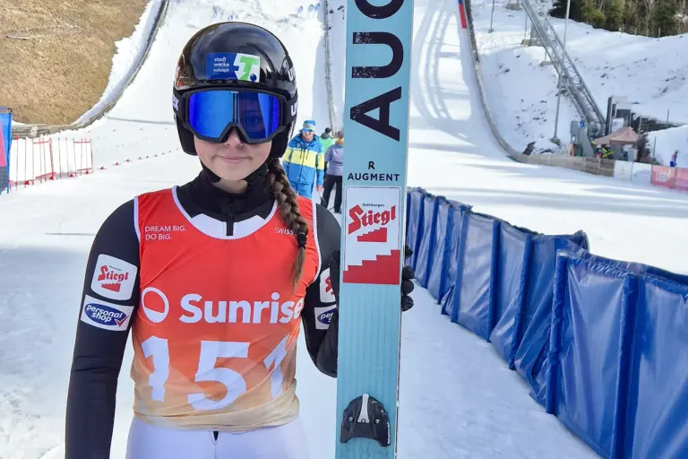 Eine junge Frau (Anna Brandner) mit einem Schi in der Hand, dahinter ist eine Sprungschanze zu erkennen.