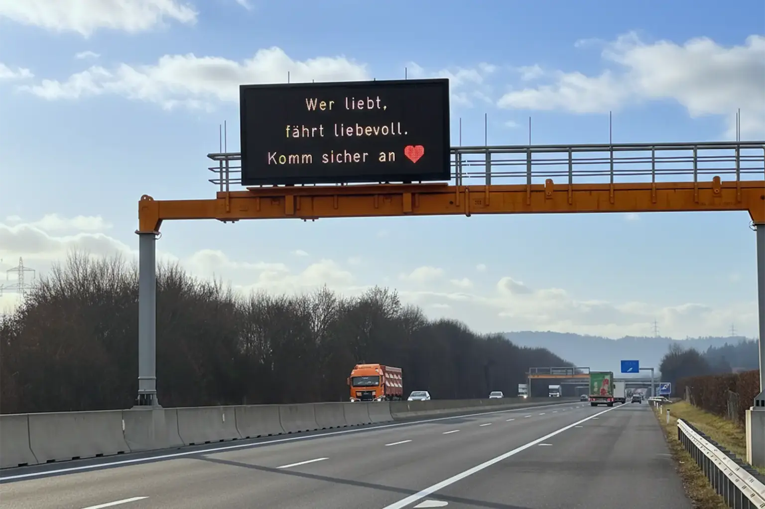 Ein großes Überkopfschild an einer Autobahn, darauf ein rotes Herz und der Text "Wer liebt, fährt liebevoll. Komm sicher an"