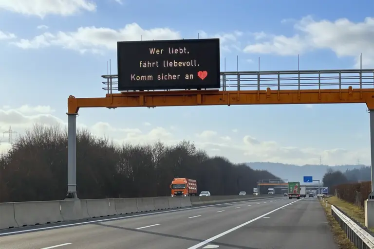 Ein großes Überkopfschild an einer Autobahn, darauf ein rotes Herz und der Text "Wer liebt, fährt liebevoll. Komm sicher an"