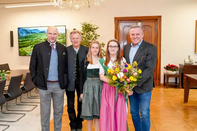 Zwei Frauen in Tracht und drei Männer in einem großen Büroraum, ein Blumenstrauß wird überreicht