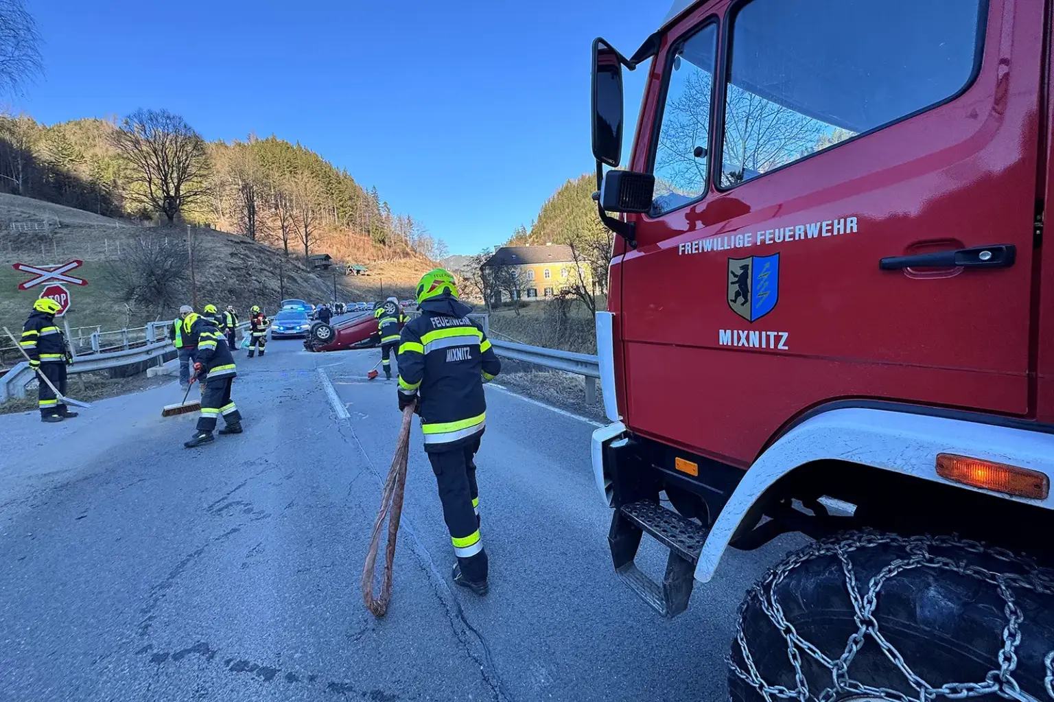Ein auf dem Dach liegendes Unfallfahrzeug auf einer Landesstraße im Bereich einer Brücke. Feuerwehrleute sichern und reinigen die Unfallstelle, am Bildrand die Fahrerkabine eines Einsatzfahrzeugs mit der Aufschrift "Freiwillige Feuerwehr Mixnitz".