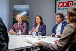 Eine Frau und zwei Männer sitzen an einem Tisch, ihnen gegenüber Zuhörerinnen