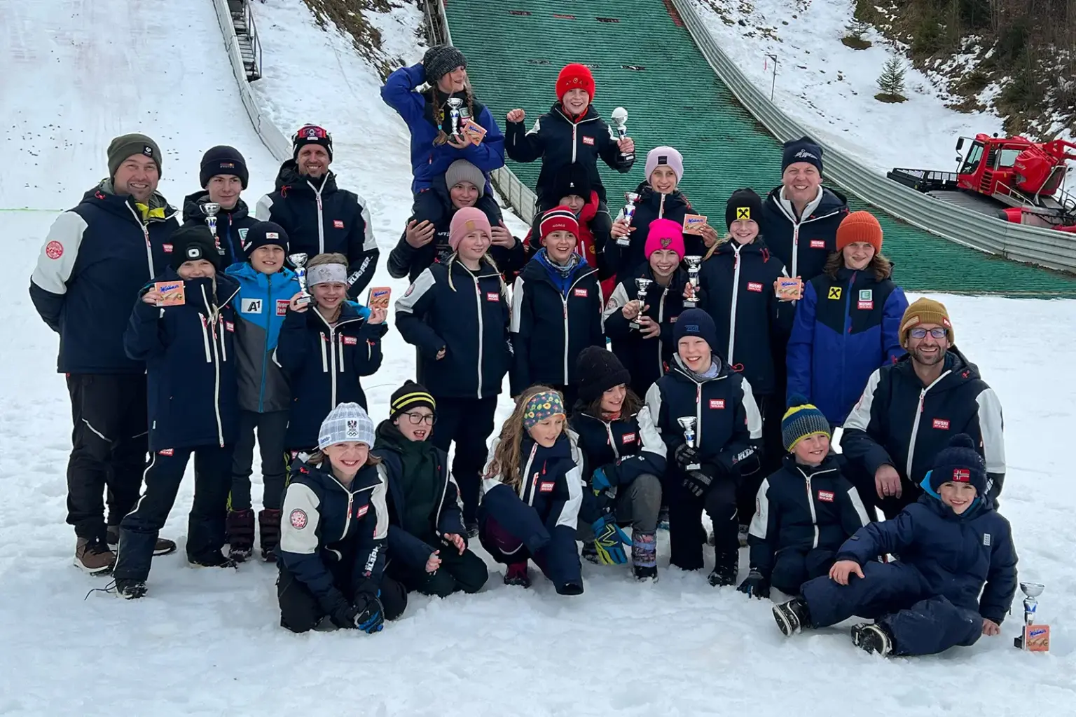Landescup der Schispringer in Mürzzuschlag Ein Gruppe von jungen Menschen in Schianzügen mit Medaillen und Pokalen in der Hand. Im Hintergrund ist eine Schisprungschanze zu erkennen.