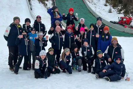 Ein Gruppe von jungen Menschen in Schianzügen mit Medaillen und Pokalen in der Hand. Im Hintergrund ist eine Schisprungschanze zu erkennen.