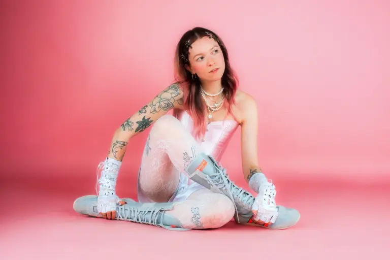 Nicht laut, aber stark Eine junge Frau mit mehreren Tätowierungen sitzt in Korsage, Strumpfhose, Boxerstiefeln und fingerlosen Handschuhen vor rosa Hintergrund