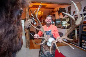 Ein bärtiger Mann in einem Raum mit Jagdtrophäen an den Wänden, in Händen hält er ein kapitales Hirschgeweih