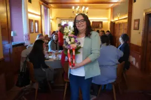Eine Frau steht im Raum mit Blumen in der Hand, im Hintergrund sind ihre Gäste beim Frühstücken.