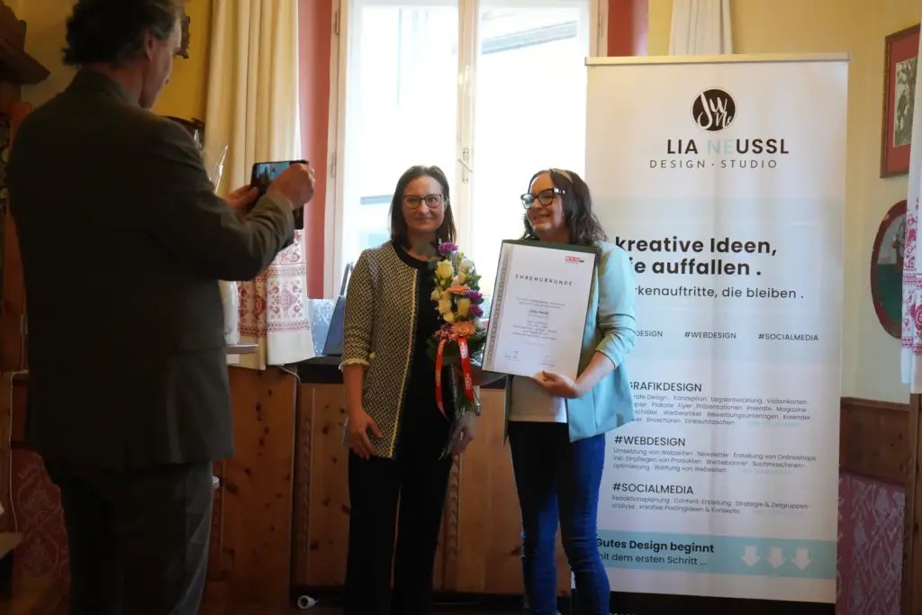 Zwei Frauen,eine hält eine Urkunde, die andere Blumen in der Hand. Dabei werden sie von einem Mann mit dem Handy fotografiert.