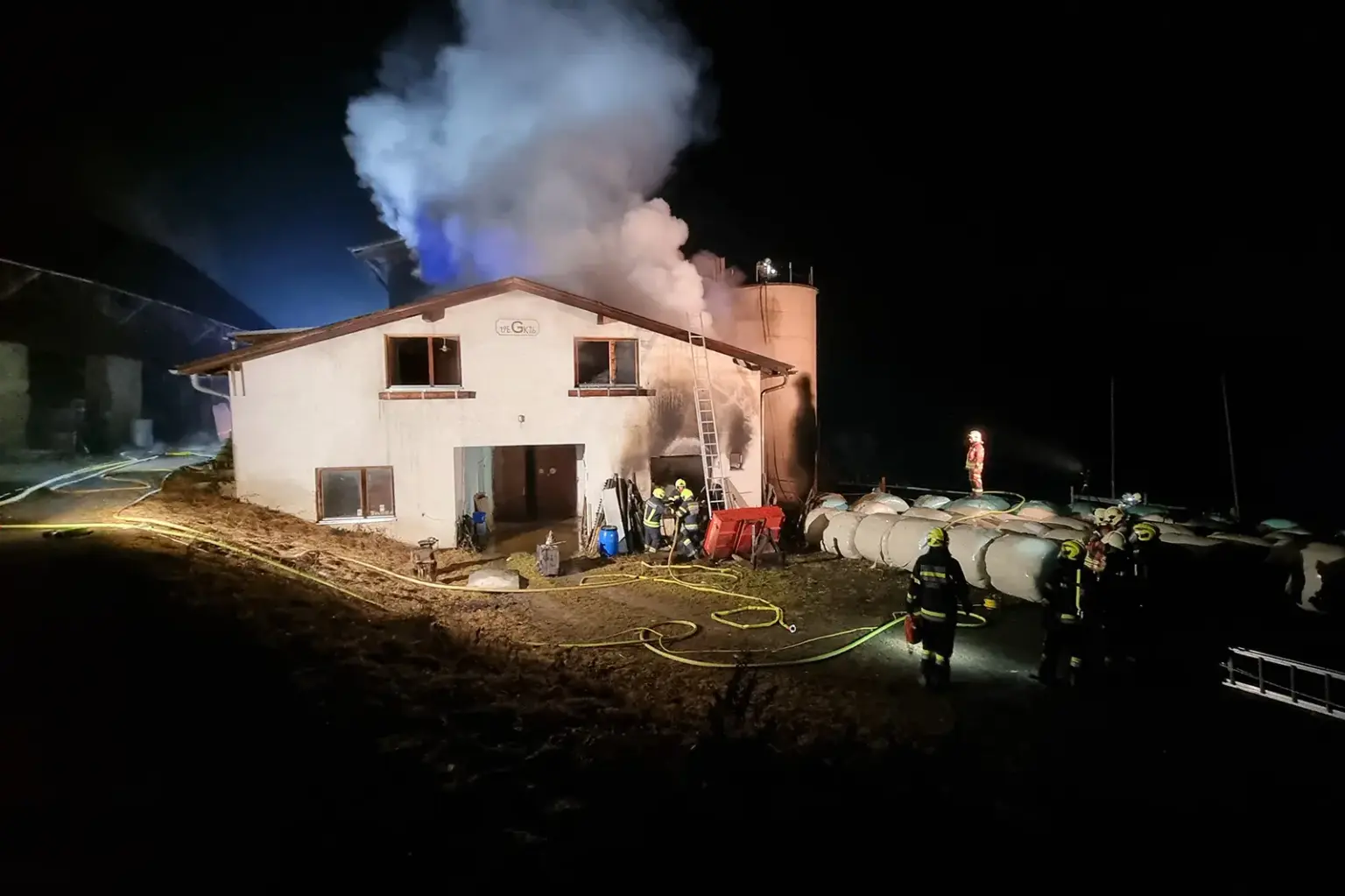 Ein Haus, über welche eine große Rauchwolke zu sehen ist. Dafür stehen einige Feuerwehrmänner.