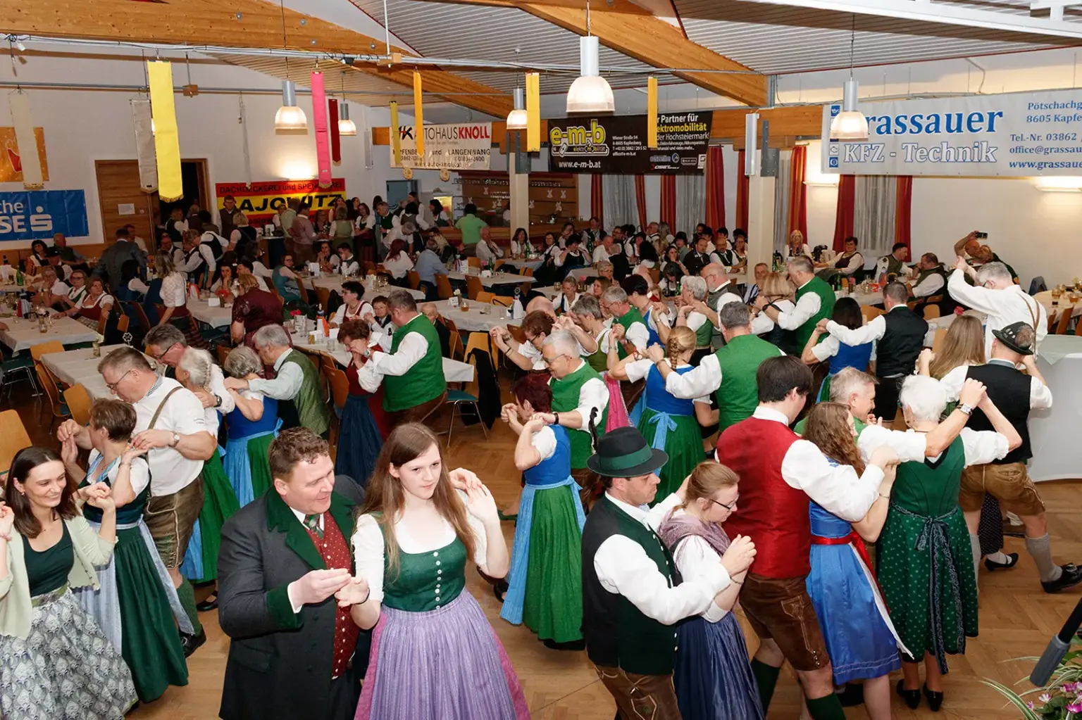 Viele Personen in Tracht tanzen oder sitzen an Tischen in einem großen Saal