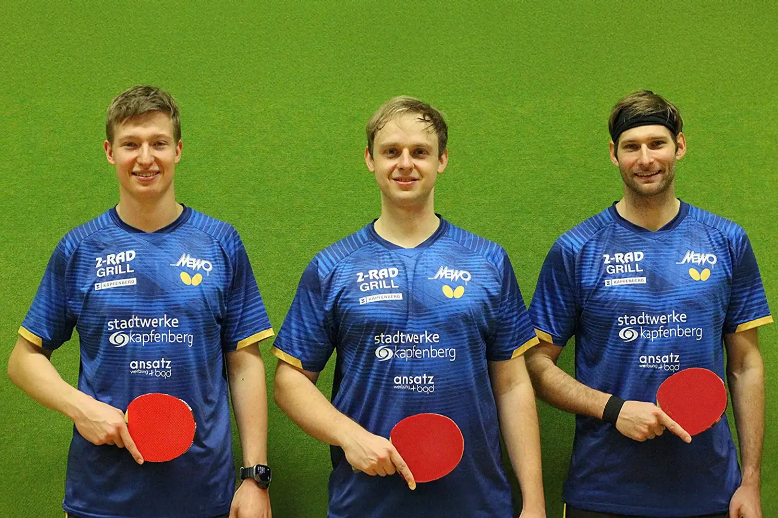 Drei junge Männer vor einer grünen Wand mit je einem Tischtennisschläger in der Hand.