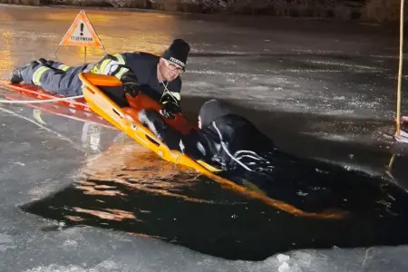 Ein auf einer zugefrorenen Wasserfläche auf dem Bauch liegender Mann in Feuerwehrkleidung zieht einen Mann mittels einer Trage aus einem ins Eis geschnittenen Loch