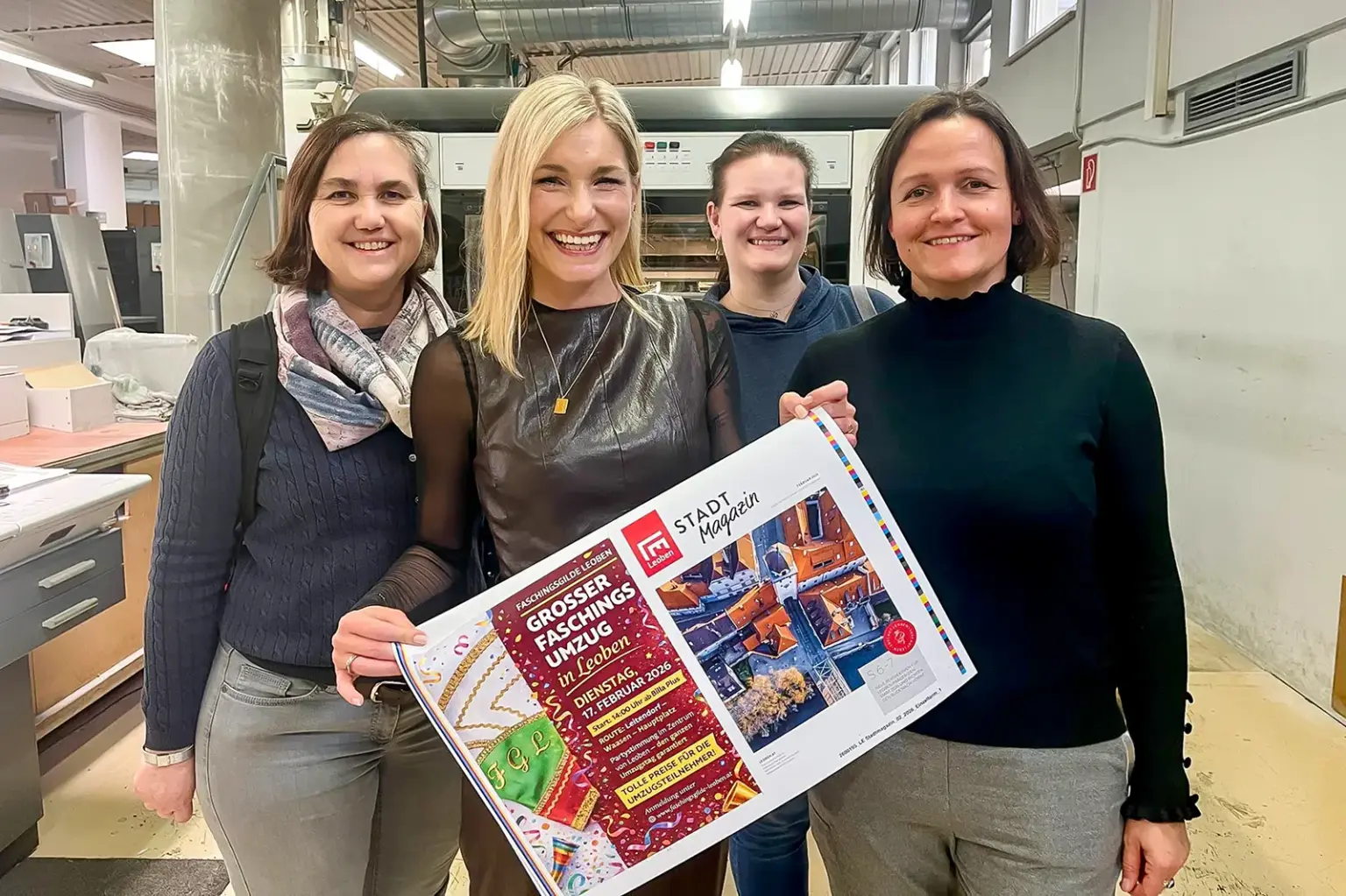 Vier lächelnde Frauen mit einer Zeitungsdoppelseite in einer Druckerei