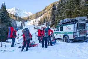 Mehrere Personen mit großen Rucksäcken stehen im Schnee, daneben sind Schi und Stöcke sowie ein Auto der Bergrettung zu erkennen.