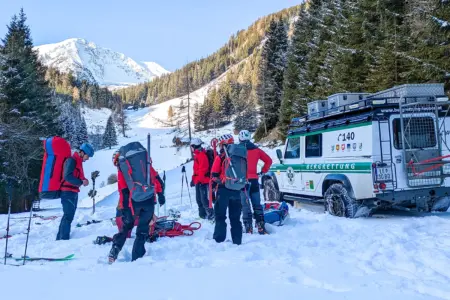 Mehrere Personen mit großen Rucksäcken stehen im Schnee, daneben sind Schi und Stöcke sowie ein Auto der Bergrettung zu erkennen.