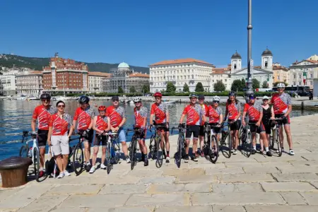 Ein Gruppe von Radfahrern steht nebeneinandern, im Hintergrund ist die Stadt Triest zu sehen.