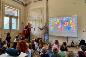 Eine Gruppe von Kindern mit einer Frau und einem Violinisten vor einem Bildschirm mit einem bunten Bild in einem alten Gebäude
