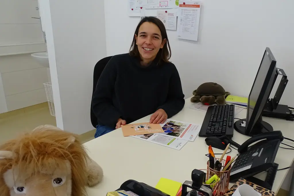 Eine Frau sitzt am Schreibtisch. Darauf steht ein Computer und ein Telefon. Zwei Stofftiere liegen am Tisch.