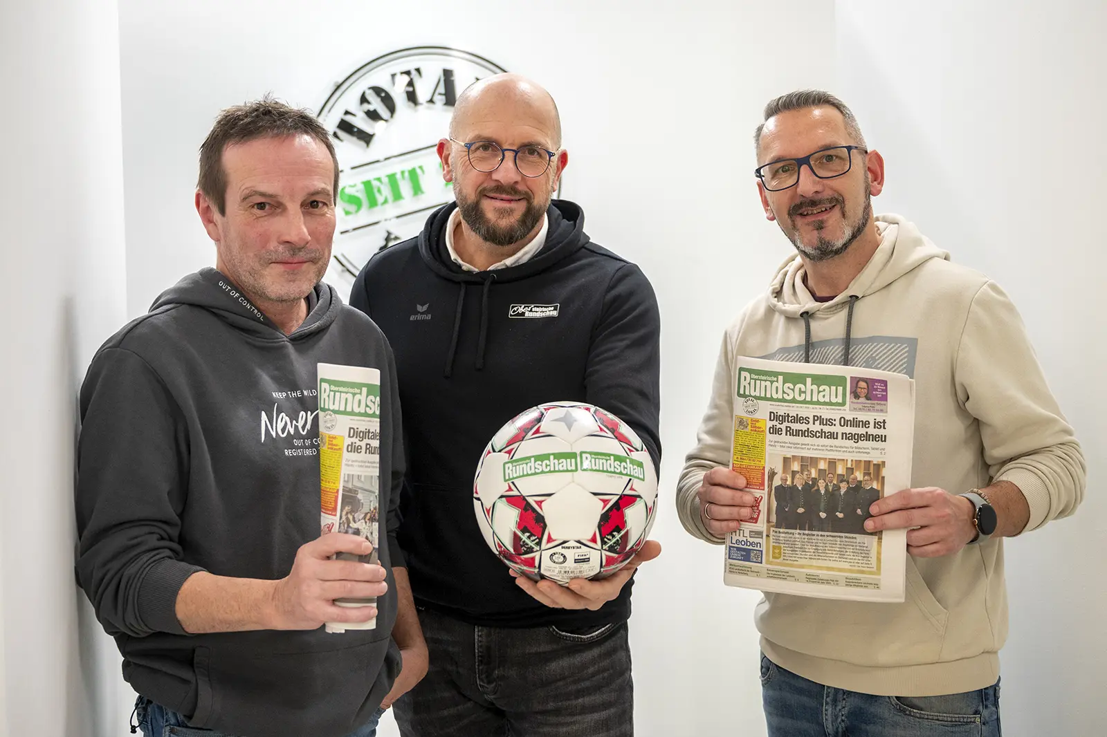 Drei Männer stehen nebeneinander, zwei haben je eine Zeitung in der Hand und einer einen Fußball.