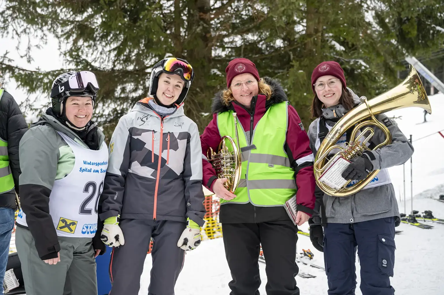 Vier Frauen stehen lächelnd im Schnee vor Tannenbäumen. Zwei tragen Skihelme und Wintersportkleidung, zwei weitere bordeauxrote Mützen und eine neongelbe Warnweste. Zwei der Frauen halten große Blasinstrumente (Tenorhorn und Tuba). Im Hintergrund sind Skipiste und Wintersport-Ausrüstung zu sehen.