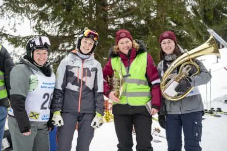 Vier Frauen stehen lächelnd im Schnee vor Tannenbäumen. Zwei tragen Skihelme und Wintersportkleidung, zwei weitere bordeauxrote Mützen und eine neongelbe Warnweste. Zwei der Frauen halten große Blasinstrumente (Tenorhorn und Tuba). Im Hintergrund sind Skipiste und Wintersport-Ausrüstung zu sehen.
