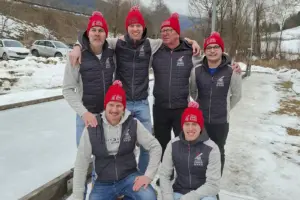 Sechs Männer stehen vor einer Eisbahn, alle sechs haben die selbe graue Jacke und rote Haube auf.