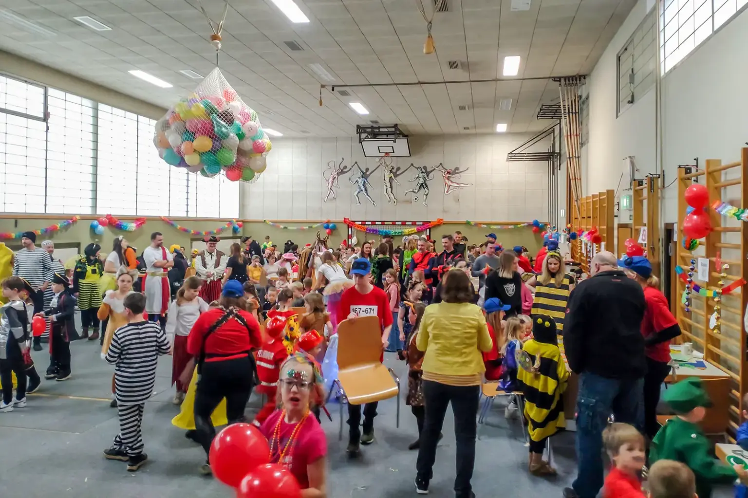 Verkleidete Kinder bei einem Faschingsfest in einem dekorierten Schulturnsaal, an der Decke ein großes Netz voller Luftballons