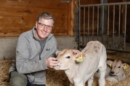 Ein Mann mit Brille kniet in einem Stall und streichelt ein Kalb