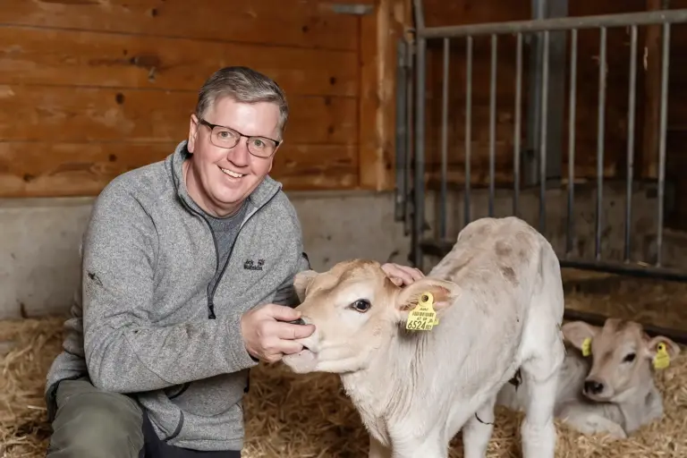 Ein Mann mit Brille kniet in einem Stall und streichelt ein Kalb