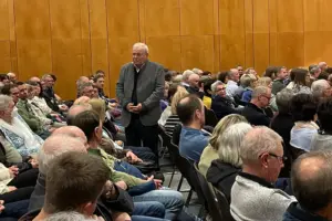 Ein älterer Mann steht inmitten von vielen sitzenden Personen